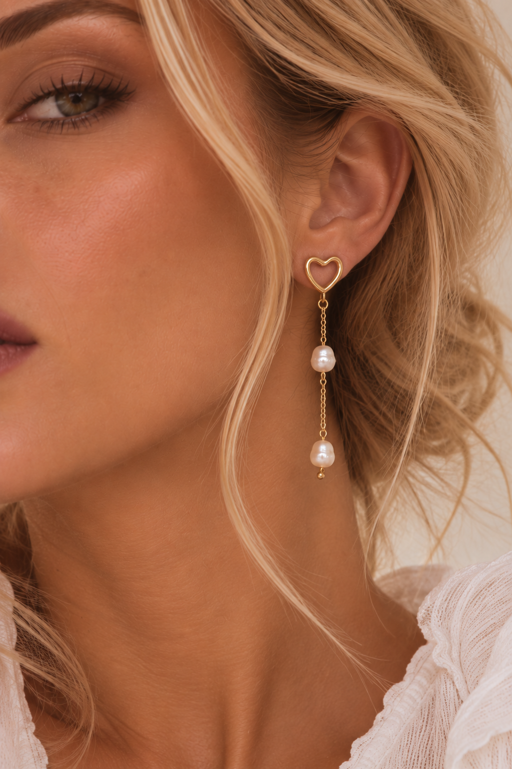 A close-up of a model's ear wearing a gold heart-shaped dangle earring with two hanging pearl beads, against a soft beige background.