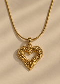 A gold heart-shaped pendant necklace with an intricate, textured openwork design, suspended from a matching gold snake chain on a warm beige fabric background.