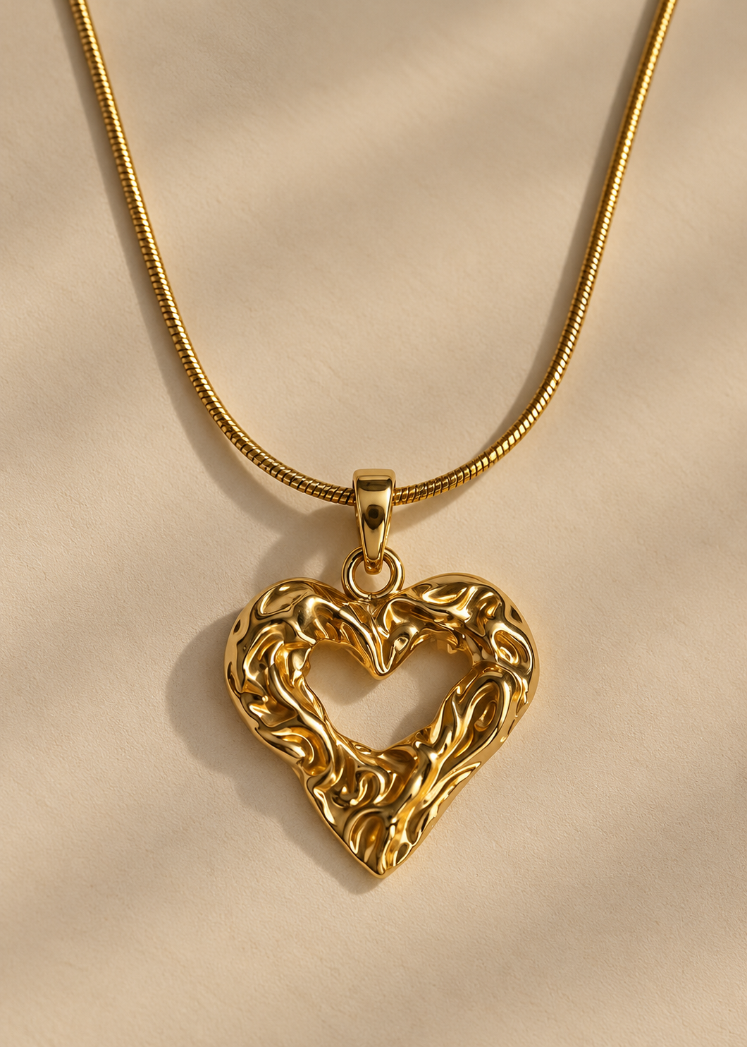 A gold heart-shaped pendant necklace with an intricate, textured openwork design, suspended from a matching gold snake chain on a warm beige fabric background.