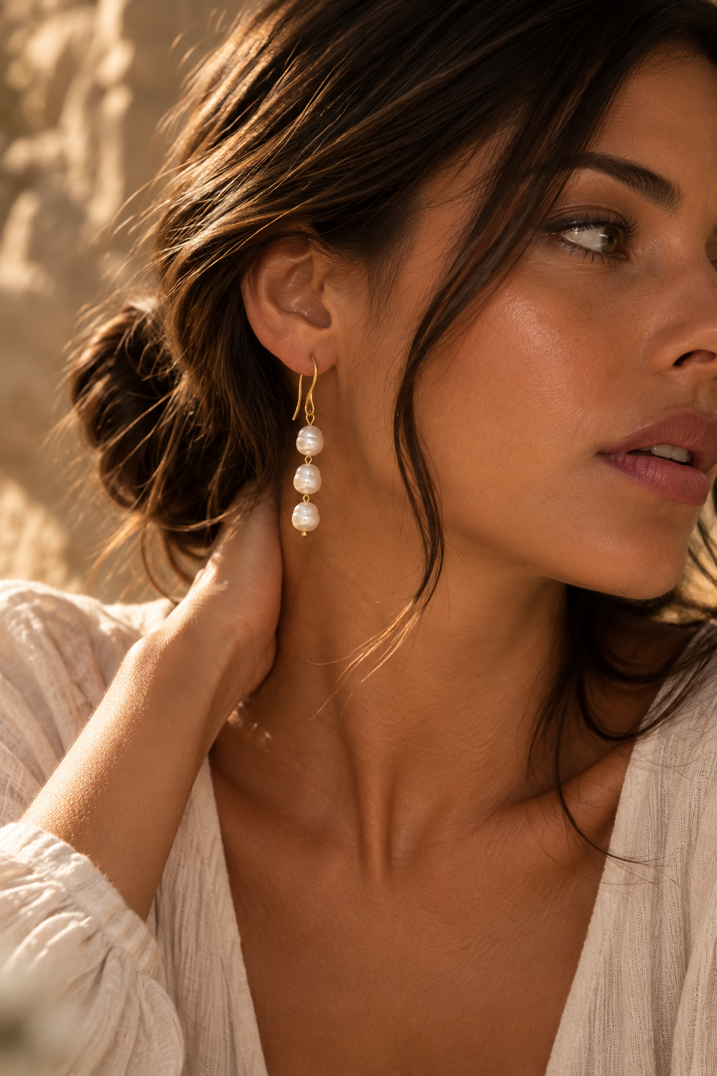 A close-up lifestyle shot of a woman wearing a gold dangle earring with three white pearl-like beads, set against a warm, sunlit neutral background.