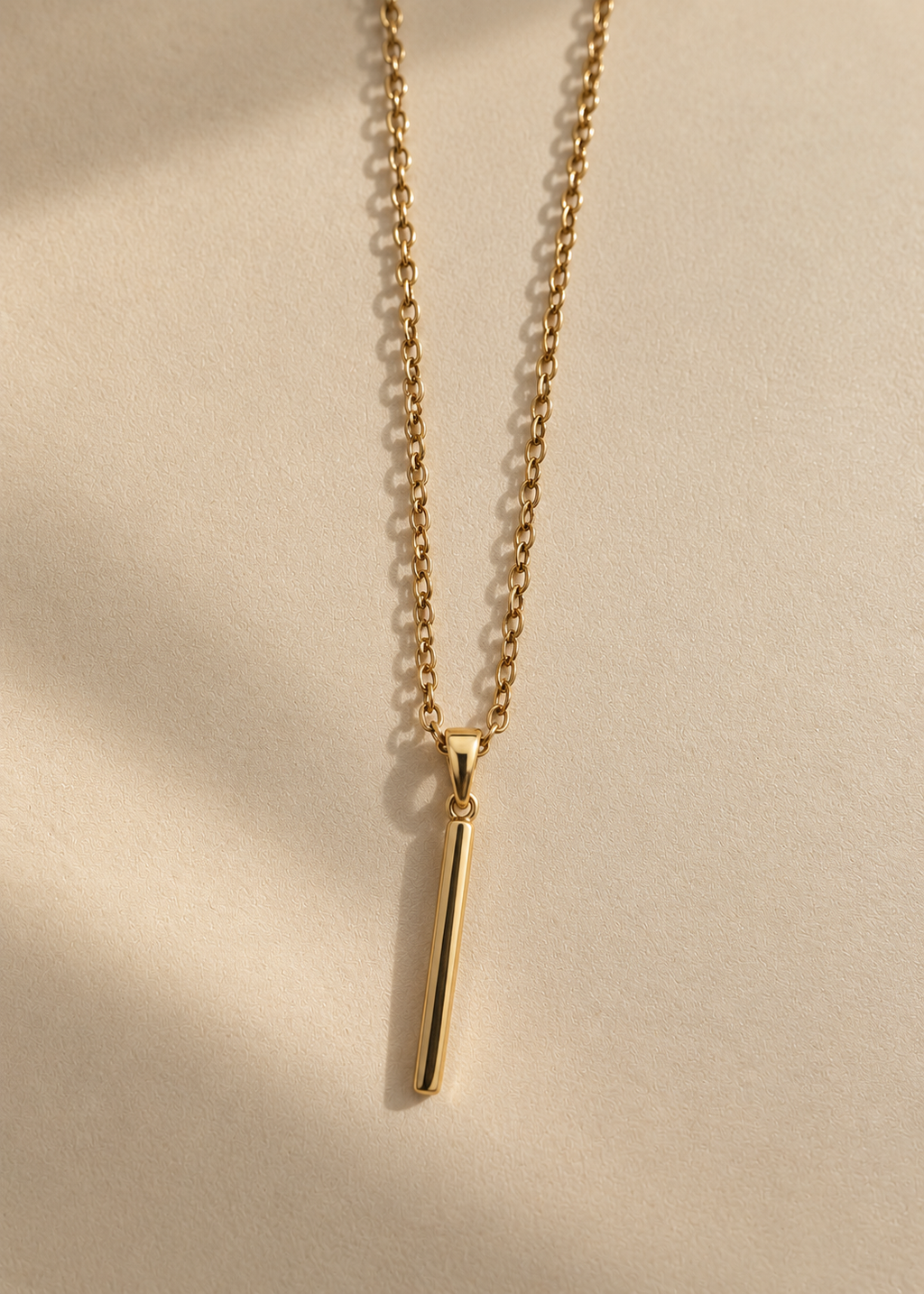 A delicate gold chain necklace with a minimalist, polished vertical bar pendant, displayed on a warm beige textured surface with soft shadows.