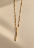 A delicate gold chain necklace with a minimalist, polished vertical bar pendant, displayed on a warm beige textured surface with soft shadows.