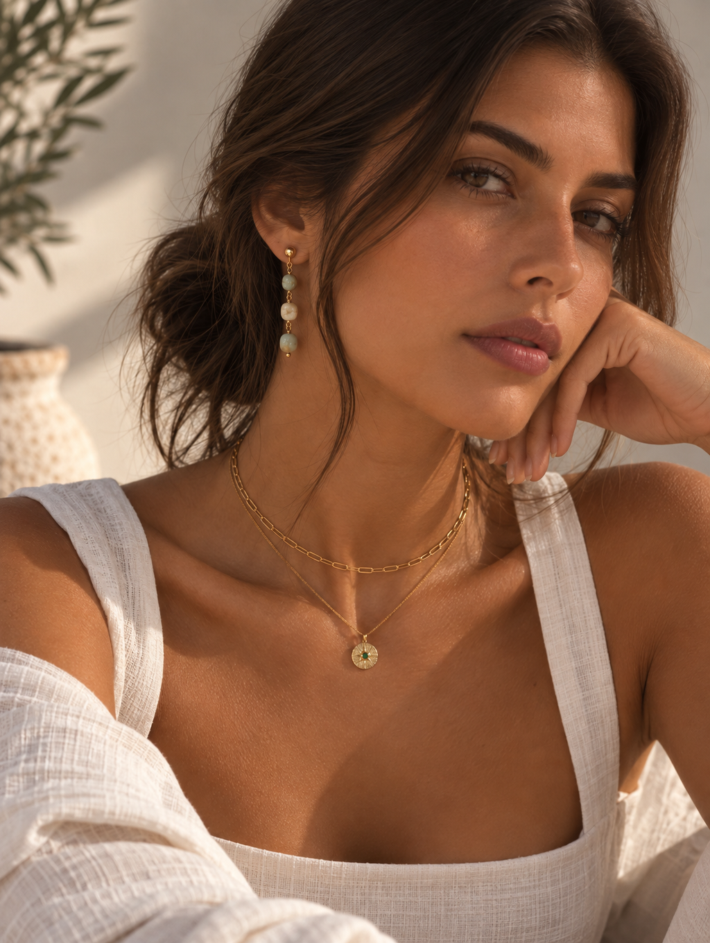 A woman wearing delicate gold jewelry, including a layered gold chain necklace with a small circular pendant and matching drop earrings, styled with a white linen top in warm natural light.