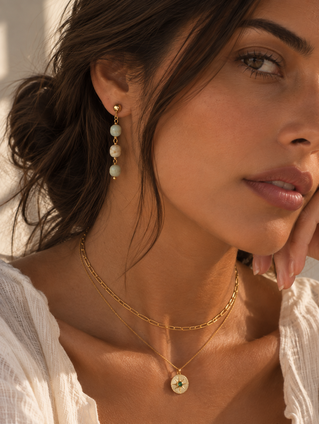 A close-up lifestyle shot of a woman wearing gold jewelry, featuring a delicate gold chain necklace with a round star pendant and a matching gold drop earring with pale green and white beads.