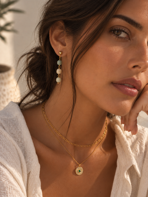 A close-up lifestyle shot of a woman wearing gold jewelry, featuring a delicate gold chain necklace with a round green and gold pendant, a gold chain necklace, and a gold drop earring with light blue and white beads.