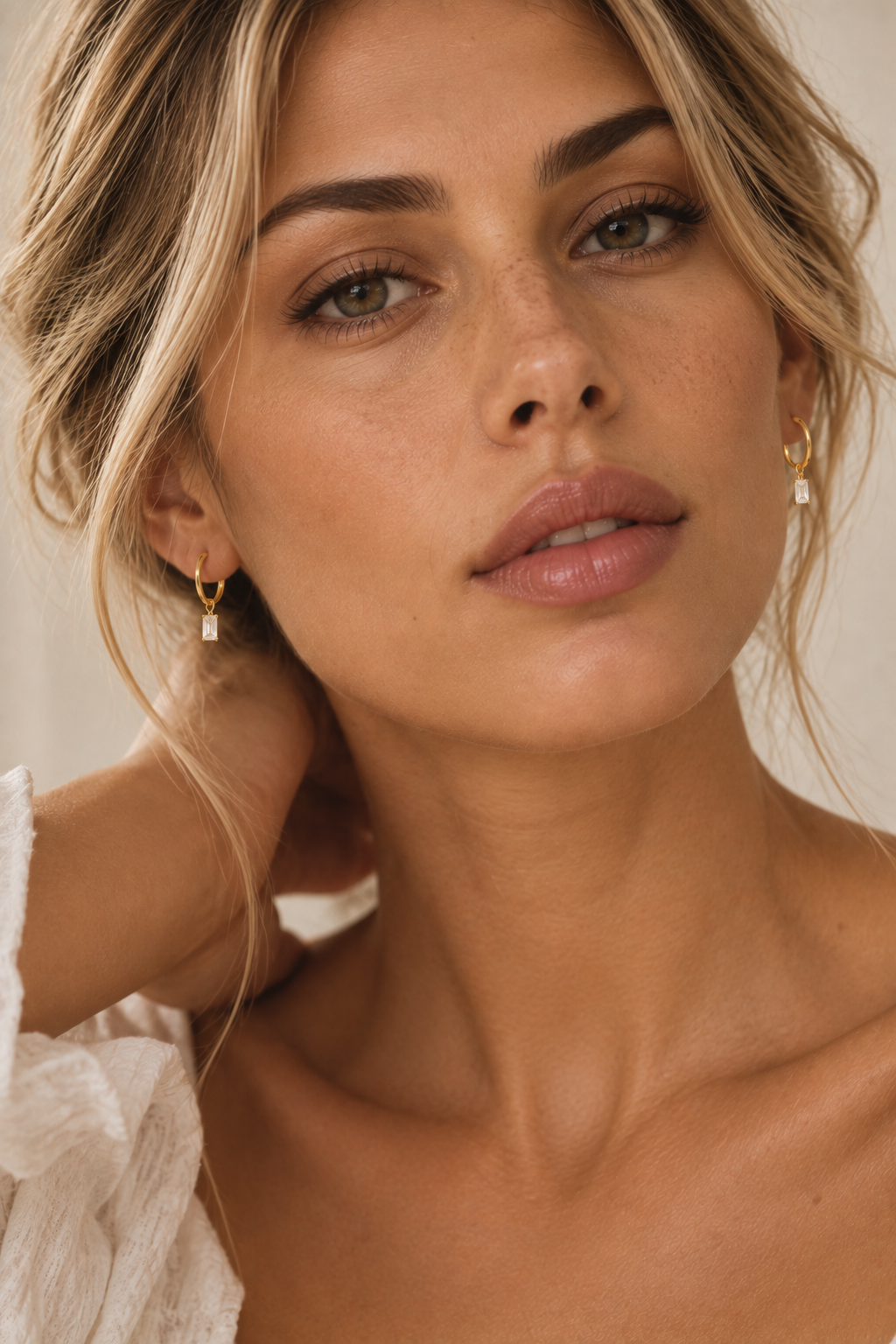 Close-up of a woman wearing small gold hoop earrings with clear rectangular drop charms, photographed against a soft beige background.