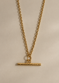 A gold chain necklace with a textured horizontal bar pendant and a small connecting ring, displayed against a plain light beige background.