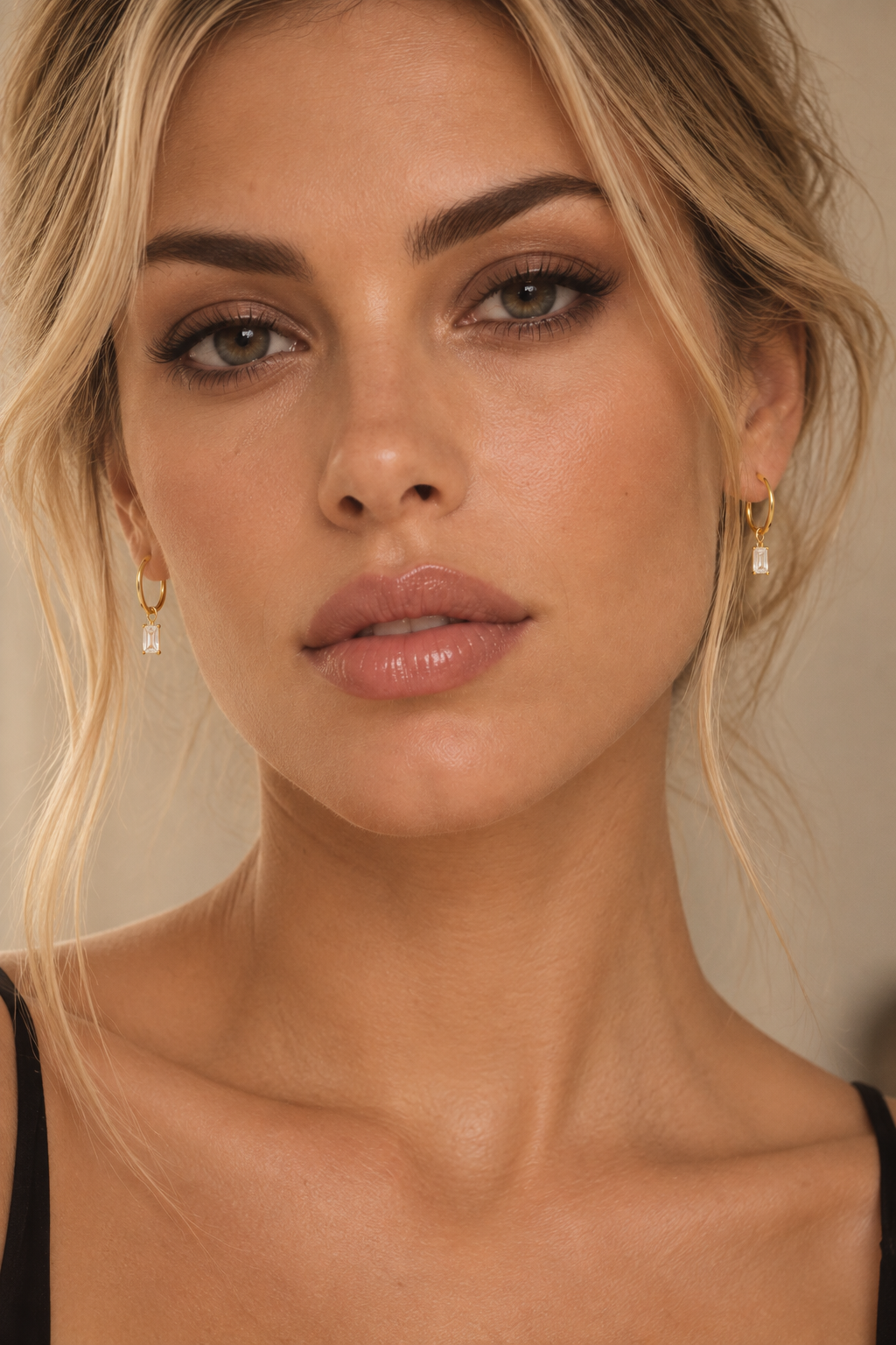 Close-up portrait of a woman wearing small gold hoop earrings with clear teardrop-shaped drops against a soft beige background.