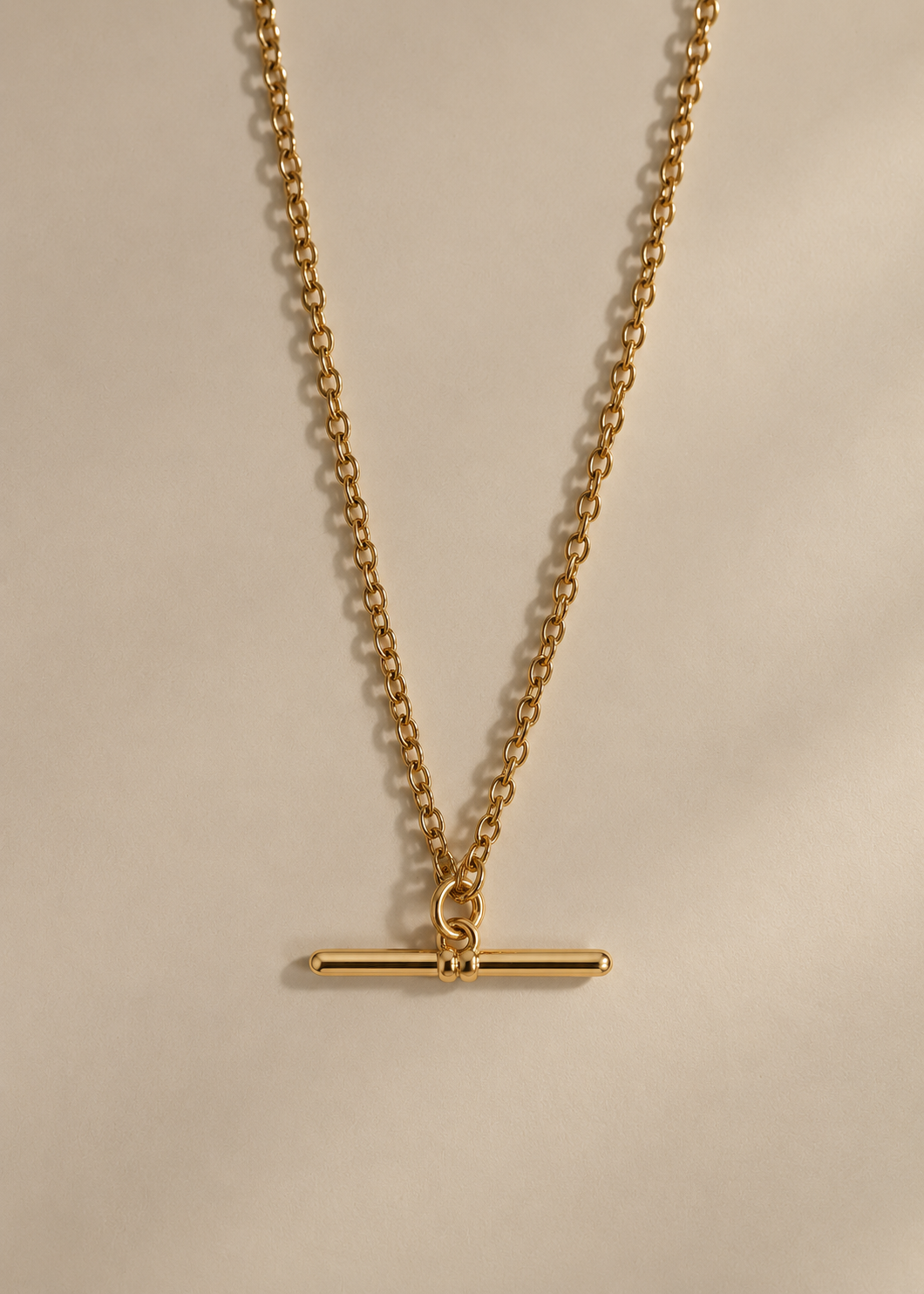 A gold chain necklace with a minimalist horizontal bar toggle pendant, displayed against a neutral beige background.