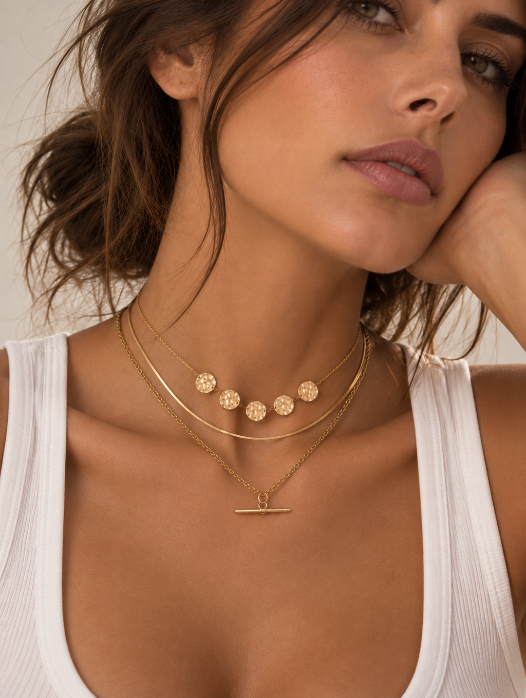 A close-up of a woman wearing a delicate gold double-layered necklace with a horizontal bar toggle pendant, styled with a white ribbed tank top.