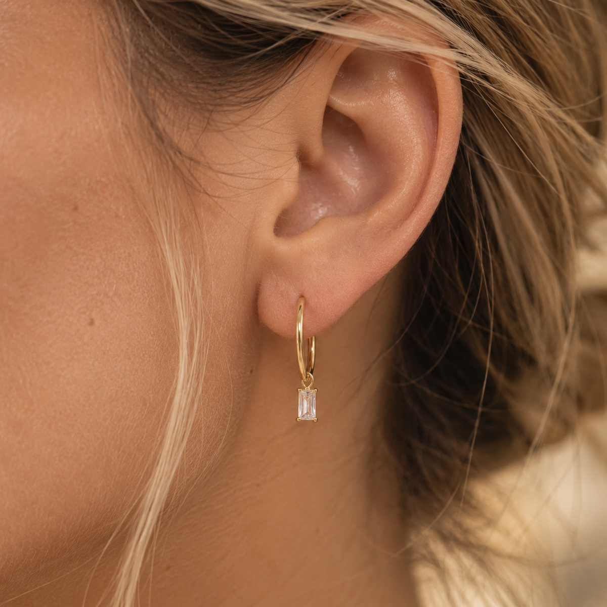 A close-up of a model's ear wearing a delicate gold huggie hoop earring with a small rectangular clear baguette gemstone dangle.