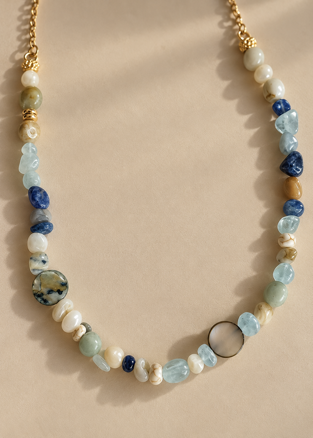 A beaded necklace with a mix of polished and textured stones in blue, green, white, and brown, accented by gold-toned metal beads and a clasp, displayed on a neutral beige background.