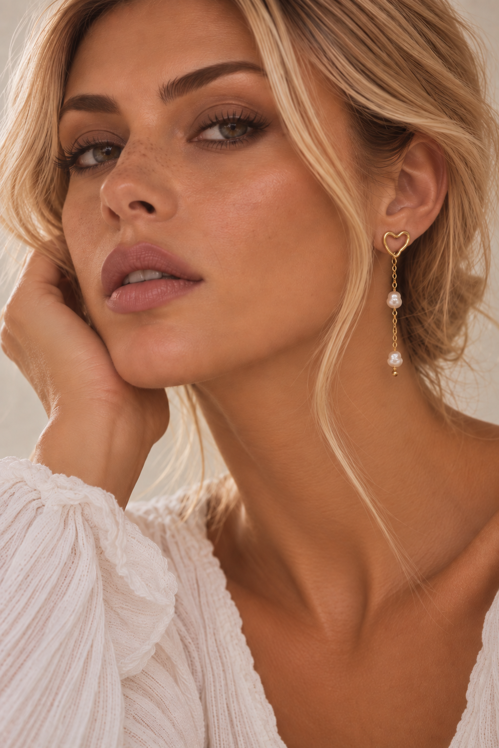 A close-up of a woman wearing a gold heart-shaped dangle earring with white pearl drops, paired with a white textured blouse against a warm beige background.