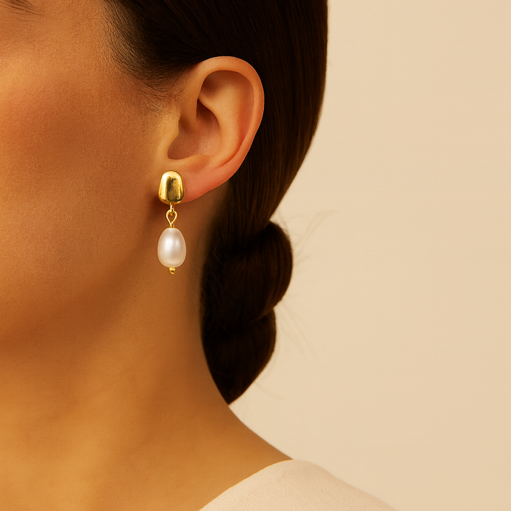Gold earring with a pearl drop worn by a person against a beige background