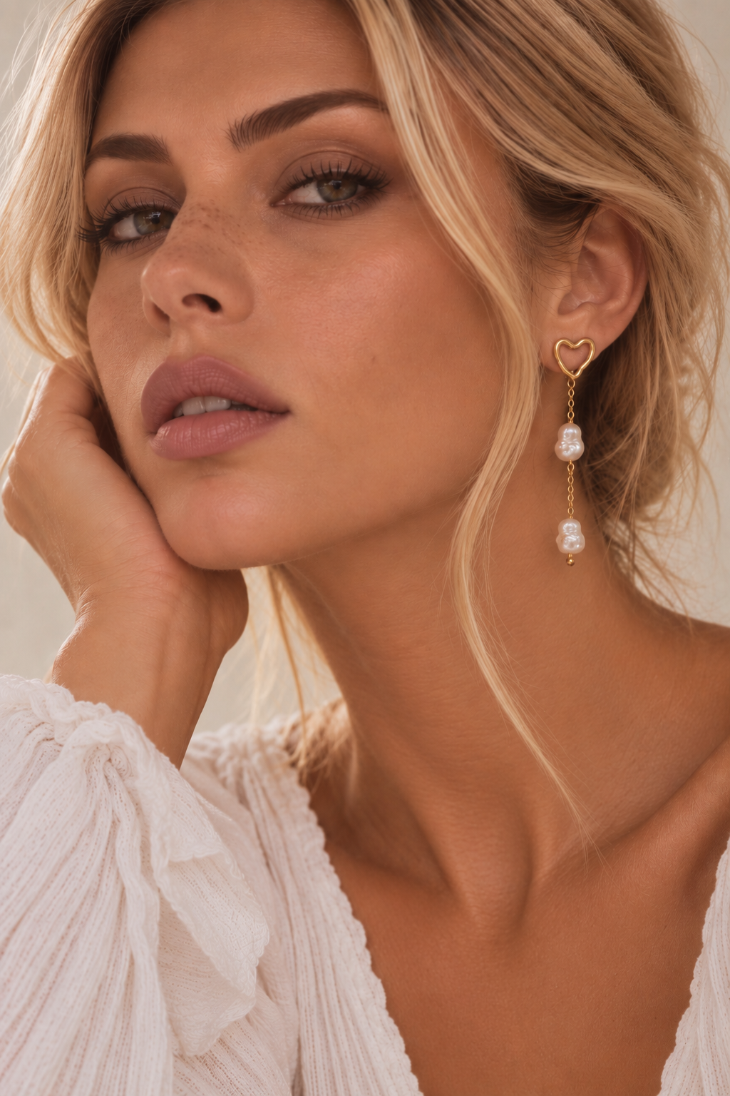 A close-up lifestyle shot of a woman wearing a gold heart-shaped dangle earring with white pearl-like beads, paired with a white ruffled blouse.