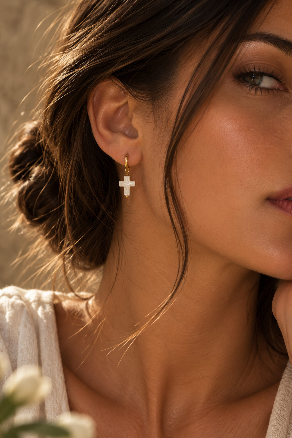 A close-up lifestyle shot of a woman wearing a small gold cross dangle earring with a white stone, set against a warm, softly blurred background.