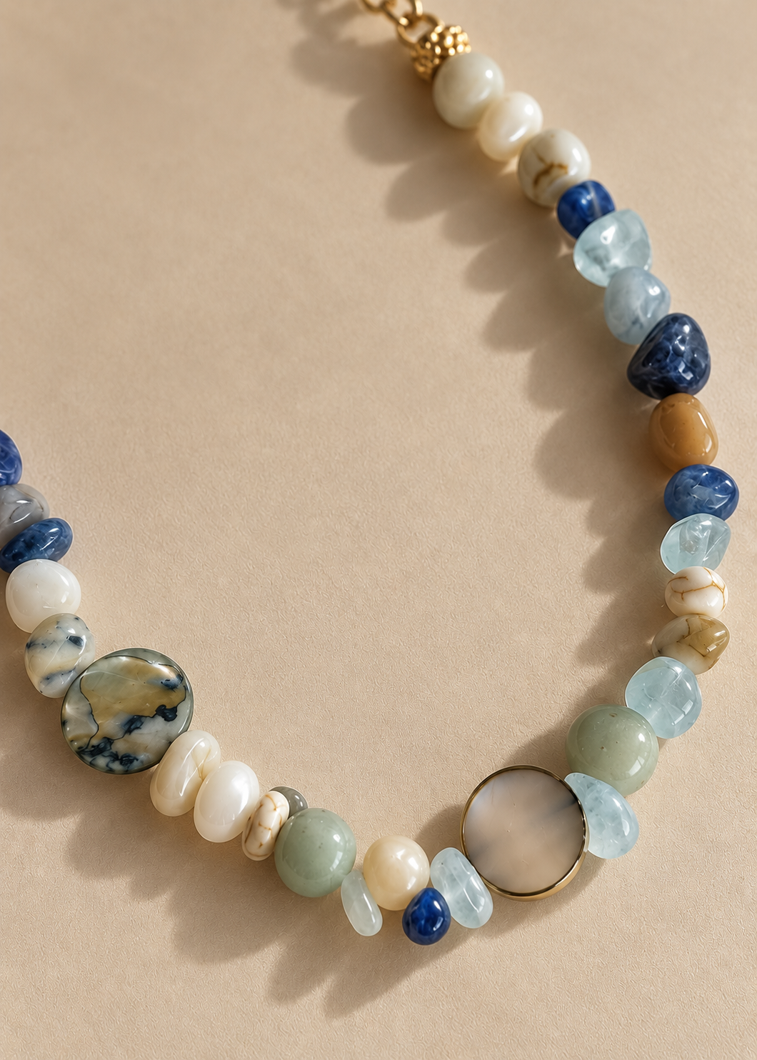 A beaded necklace with a mix of polished and textured stones in blue, green, white, and brown, accented by gold-toned metal beads and a clasp, displayed on a neutral beige background.