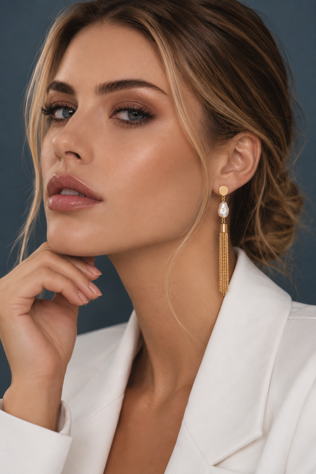 A close-up of a woman wearing a long gold dangle earring with a white teardrop-shaped stone, paired with a white blazer against a dark slate blue background.