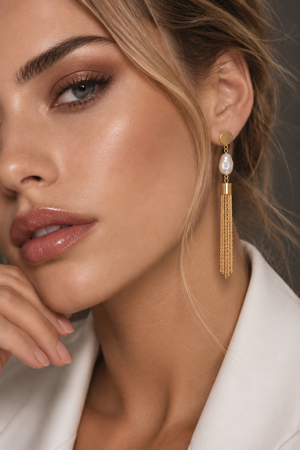A close-up of a woman wearing a long gold tassel dangle earring with a white pearl, paired with a white blazer against a dark gray background.