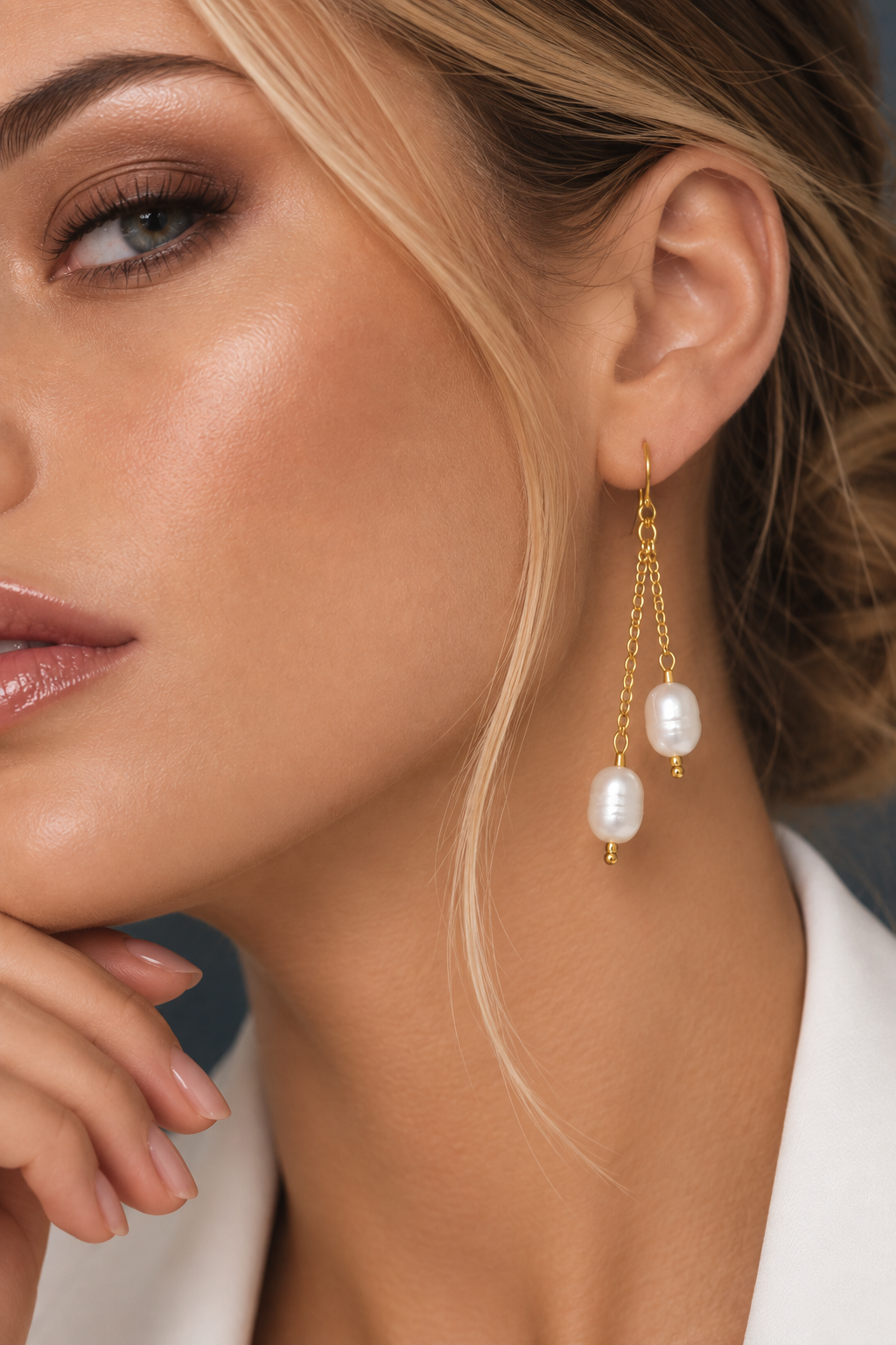 Close-up of a woman wearing a pair of gold dangle earrings with white baroque pearls, shown against her ear and neck.