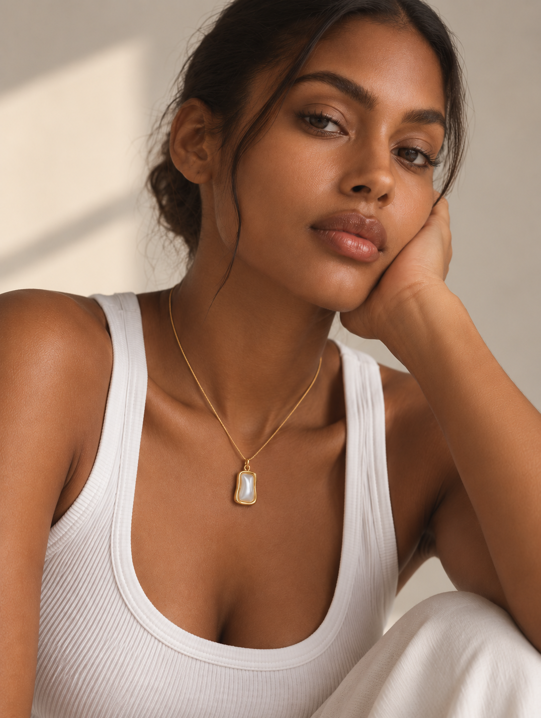 A woman wearing a delicate gold chain necklace with a teardrop-shaped white pearl pendant, styled with a white ribbed tank top against a warm, softly lit background.