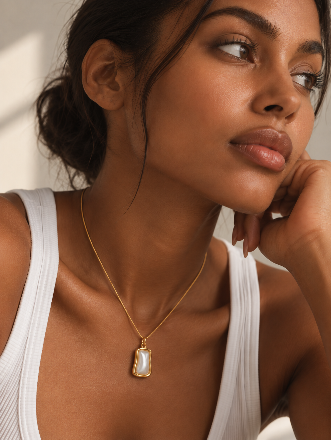 A woman wearing a delicate gold chain necklace with a teardrop-shaped white pearl pendant, styled with a white ribbed tank top against a warm, softly lit background.