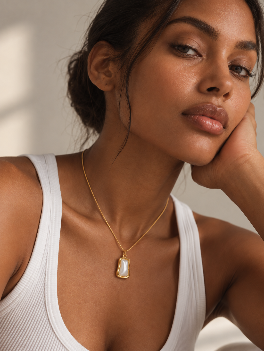 A woman wearing a delicate gold chain necklace with a teardrop-shaped white pearl pendant, styled with a white ribbed tank top against a warm, softly lit background.