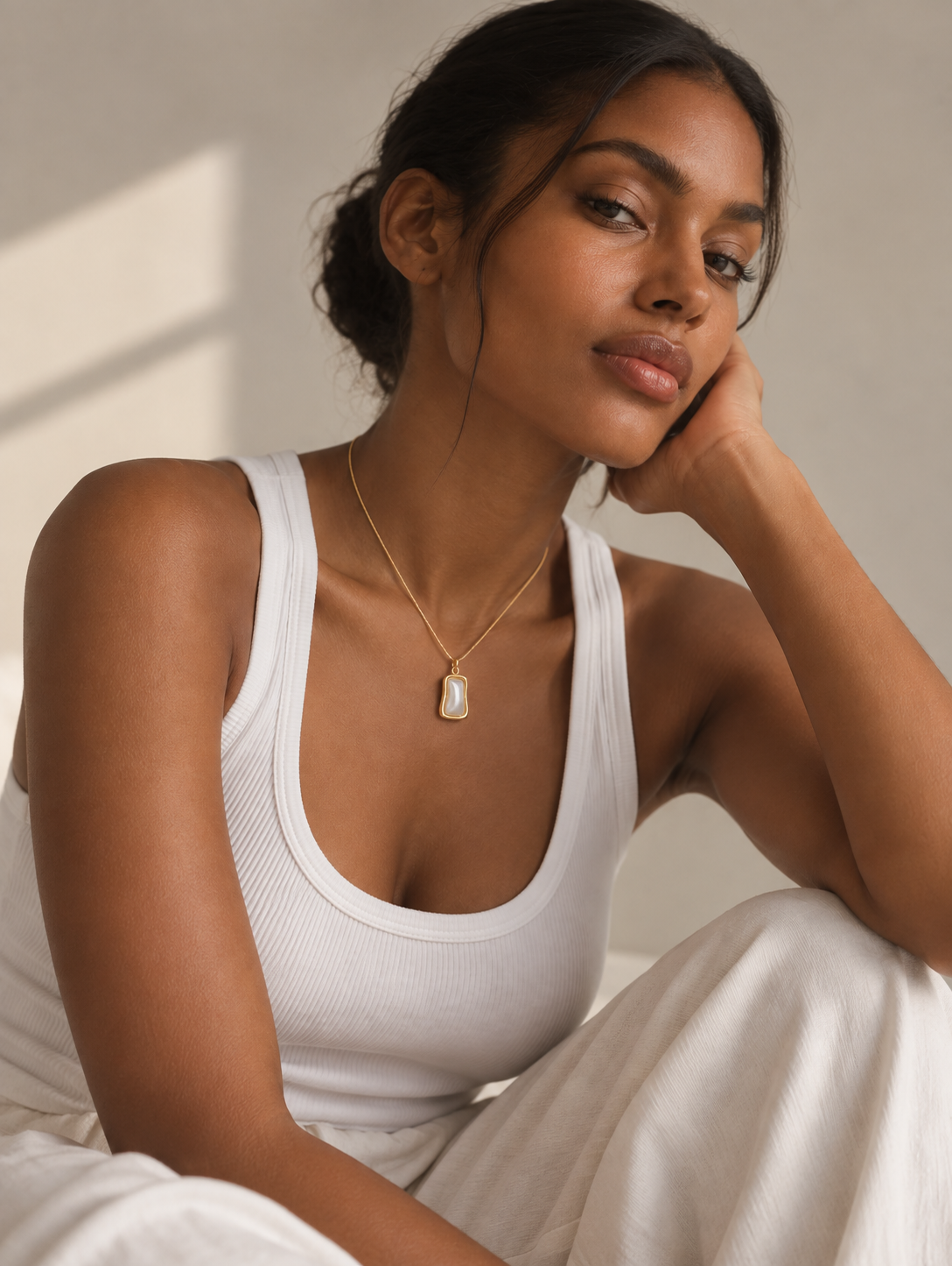 A woman wearing a delicate gold necklace with a small white rectangular pendant, styled with a white ribbed tank top and white pants in a softly lit indoor setting.