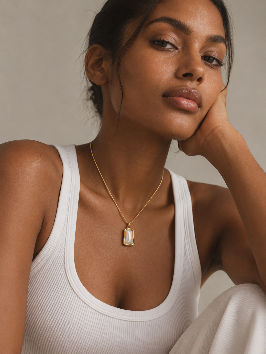 A woman wearing a delicate gold pendant necklace with a white stone, styled with a white ribbed tank top against a neutral beige background.