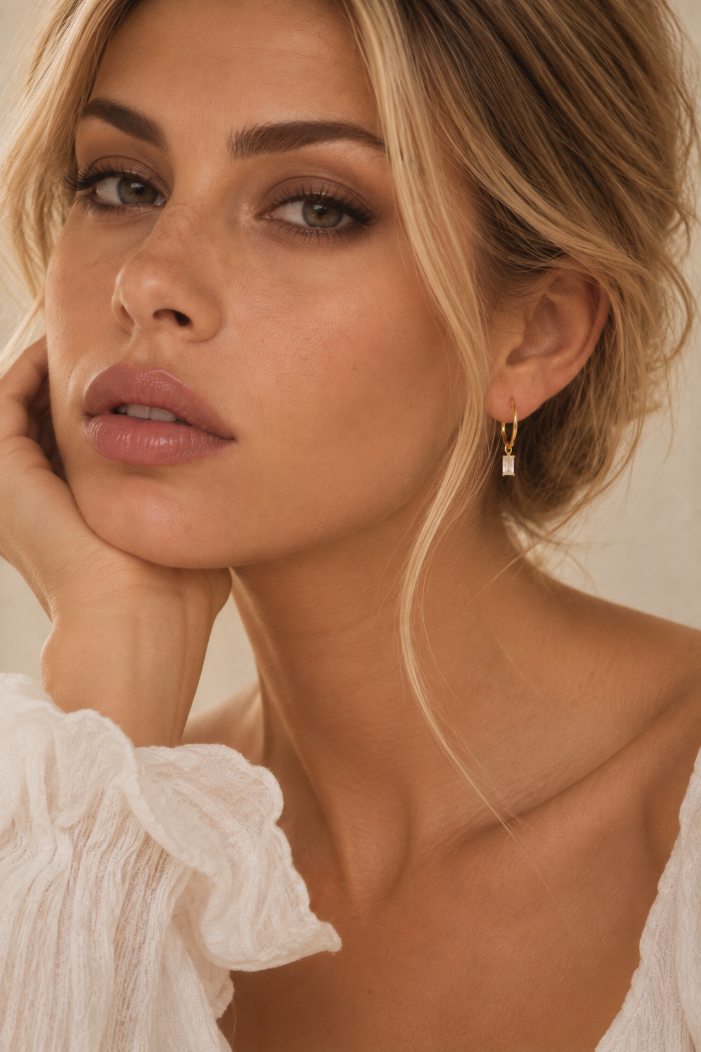 A close-up lifestyle shot of a woman with blonde hair wearing a delicate gold huggie hoop earring with a small white rectangular charm.