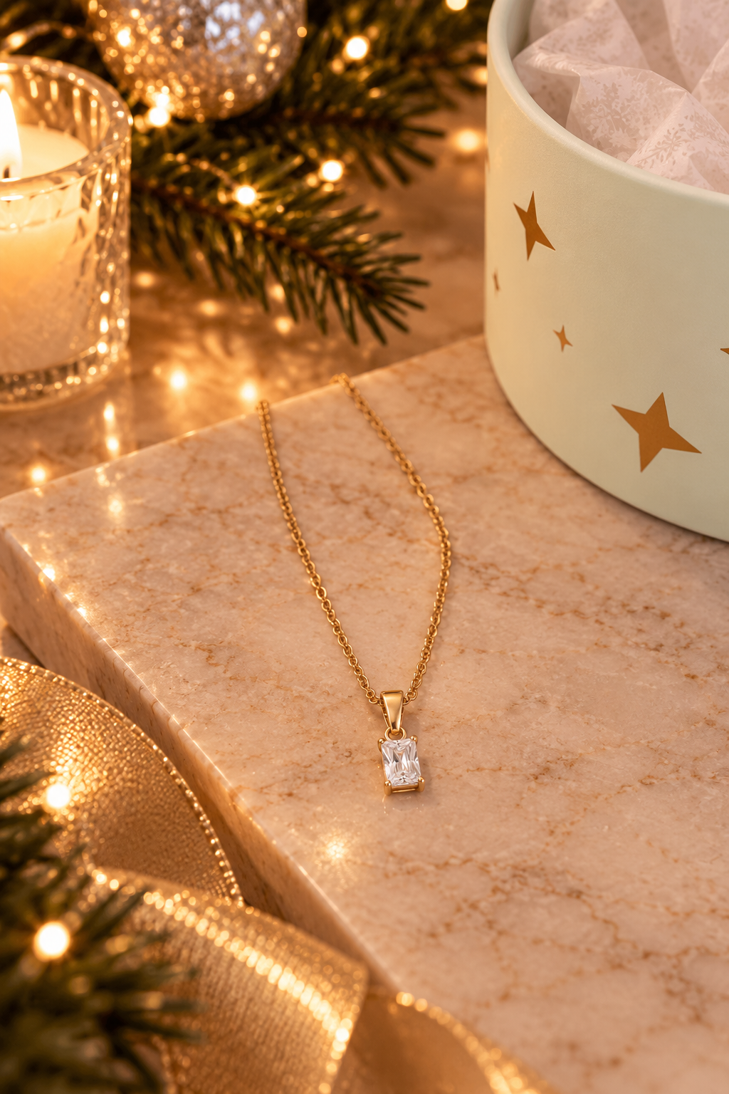 A gold necklace with a rectangular clear gemstone pendant rests on a beige marble surface, styled with warm holiday lights, pine branches, a candle, and a gift box with gold stars.