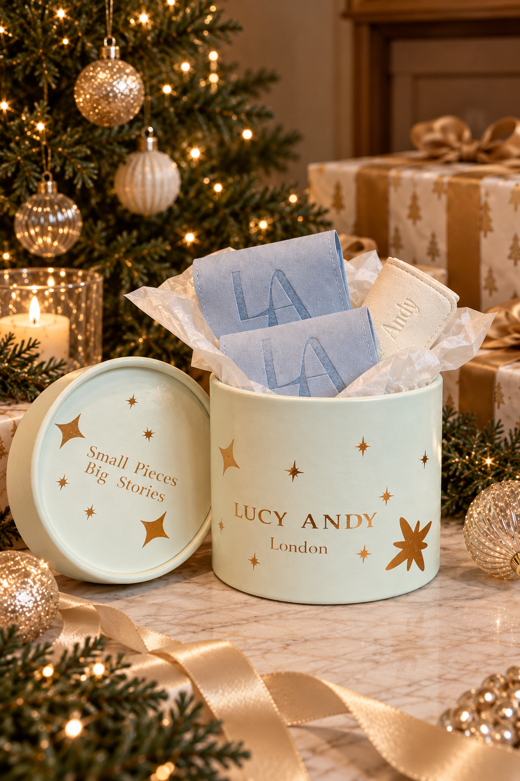 A festive Christmas-themed gift scene featuring a round pale green Lucy Andy London box with gold star accents, opened to reveal blue and cream roller velvet bags and a white jewellery  travel bag . The matching lid sits beside it on a marble surface, surrounded by a decorated Christmas tree, wrapped presents, and gold ribbons.