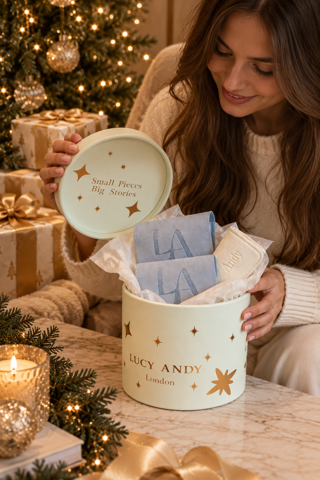 A woman in a cozy sweater opens a round pastel green gift box labeled 'LUCY ANDY London' with gold stars, revealing folded blue and white jewellery bags inside. The scene is decorated for Christmas with a lit tree, wrapped presents, and a candle in the background.