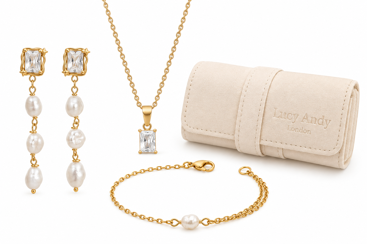 Gold jewelry set with a necklace, drop earrings, bracelet, and a cream-colored velvet pouch. The jewelry features clear gemstones and white pearls, with the pouch embossed with the brand name 'Lucy Andy London'.