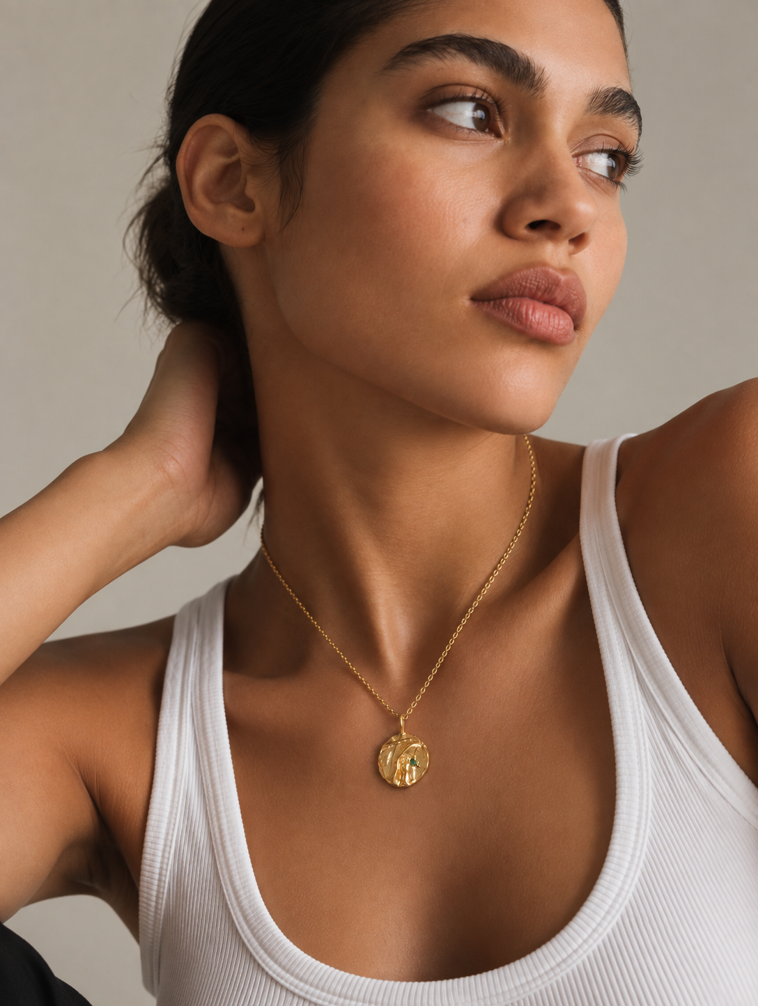 A close-up of a woman wearing a delicate gold chain necklace with a round, textured gold pendant, paired with a white ribbed tank top against a neutral beige background.