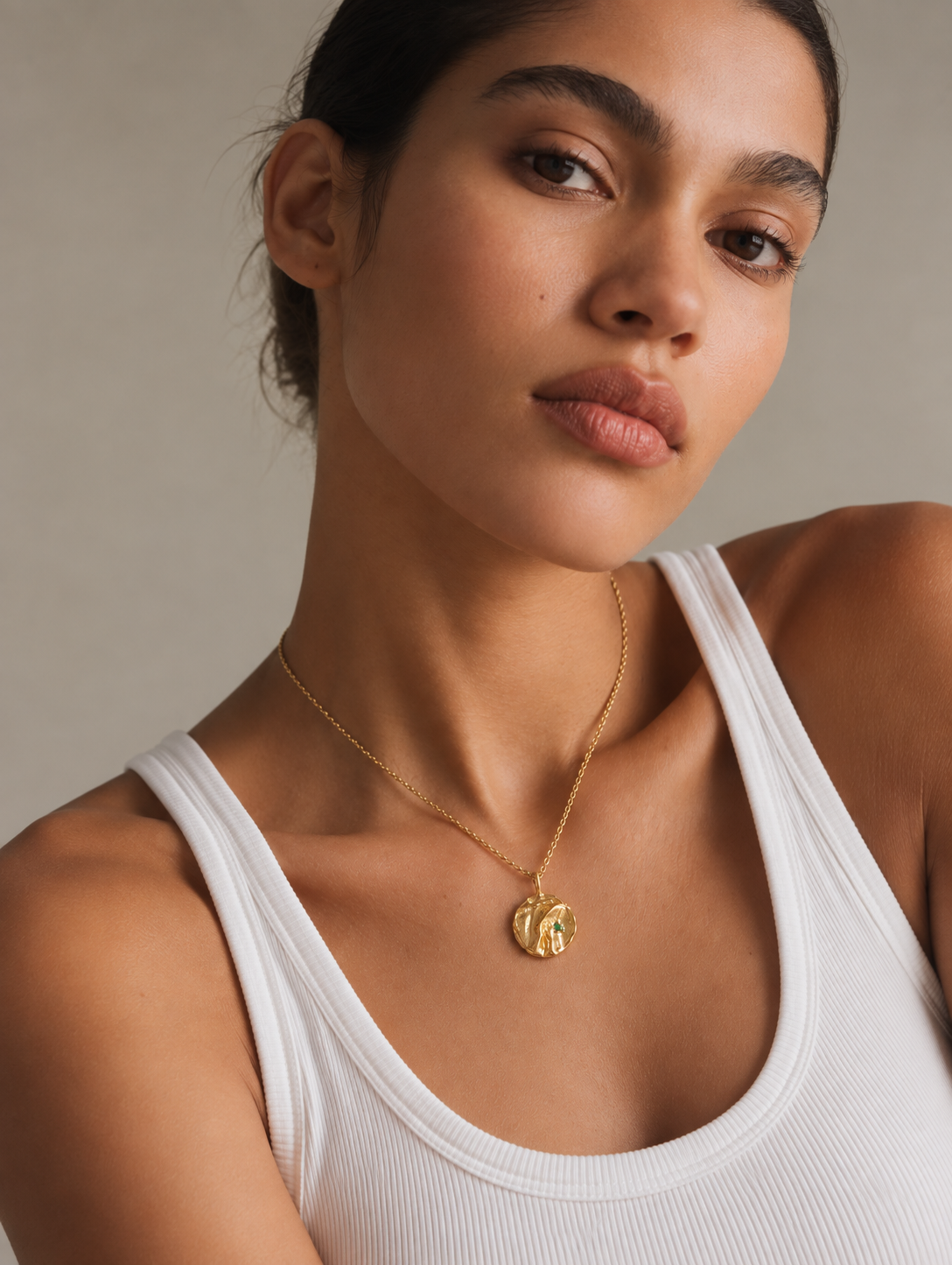 A close-up of a woman wearing a delicate gold chain necklace with a textured circular pendant, paired with a white ribbed tank top against a neutral beige background.