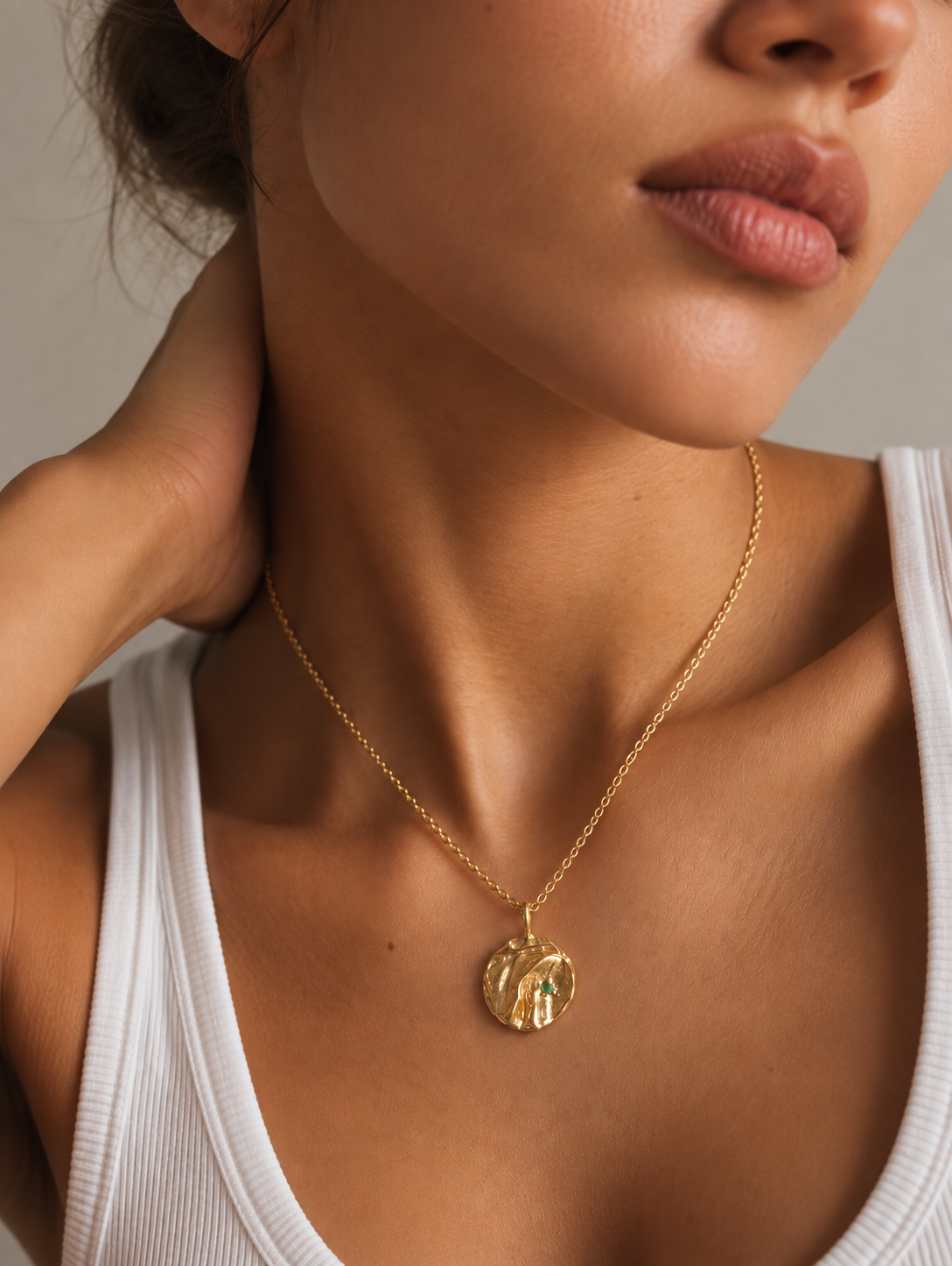 A close-up lifestyle shot of a model wearing a delicate gold chain necklace with a textured, organic-shaped gold pendant featuring a small green inlay, styled with a white ribbed tank top.