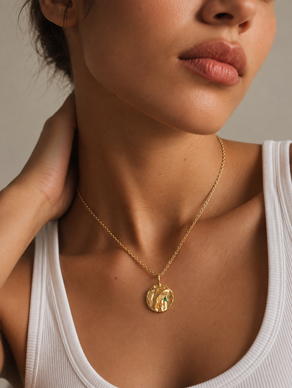 A close-up lifestyle shot of a model wearing a delicate gold chain necklace with a textured, organic-shaped gold pendant featuring a small green inlay, styled with a white ribbed tank top.