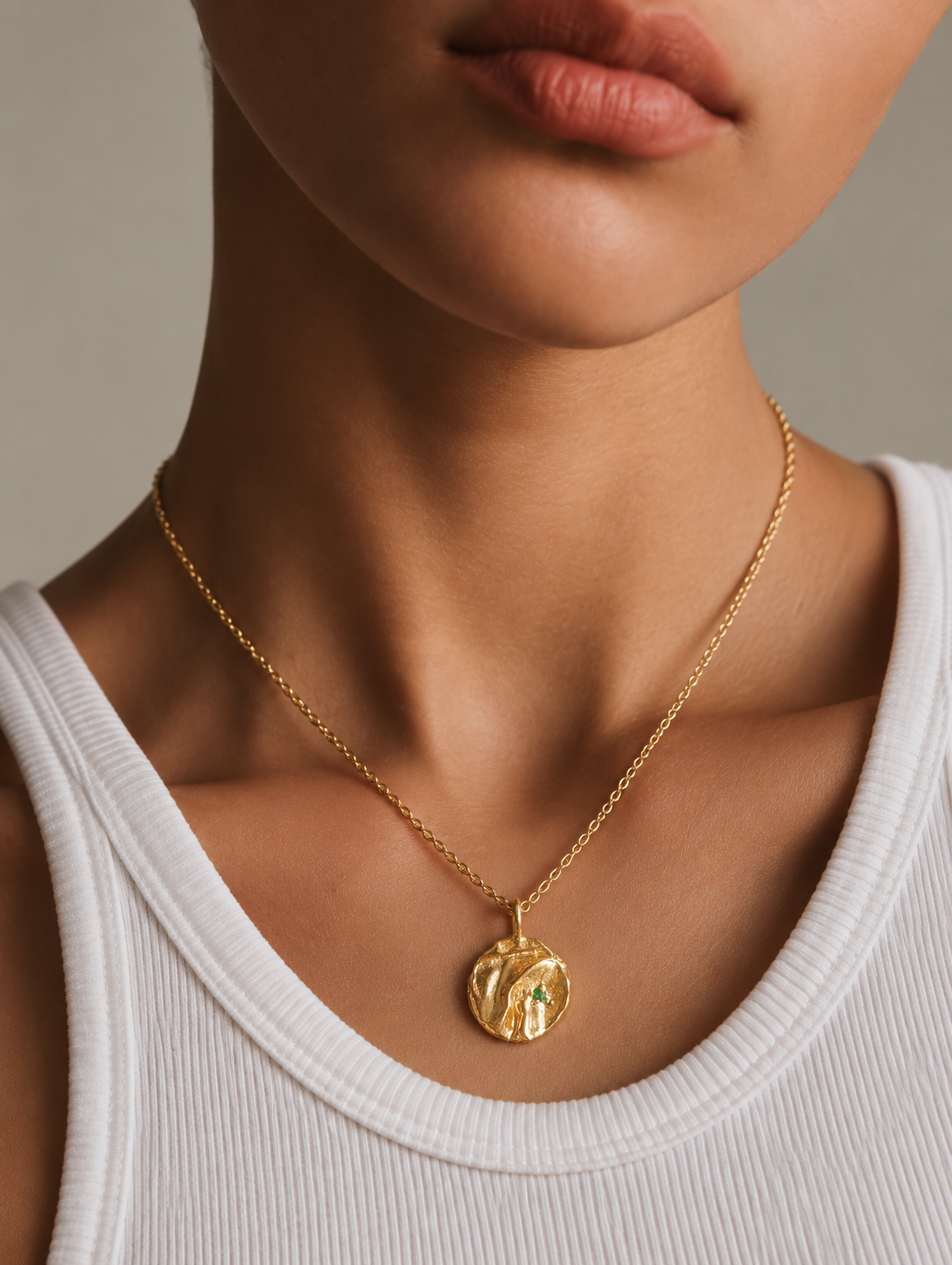 A close-up of a model wearing a delicate gold chain necklace with a textured, circular gold pendant, styled with a white ribbed tank top against a neutral gray background.