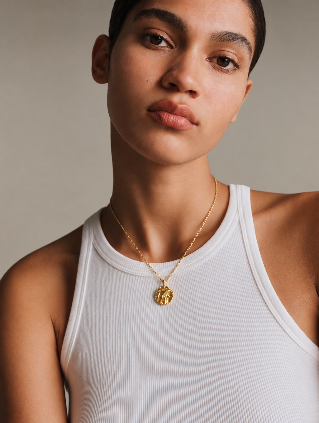 A close-up of a model wearing a delicate gold chain necklace with a small, textured gold medallion pendant, paired with a white ribbed tank top against a neutral beige background.