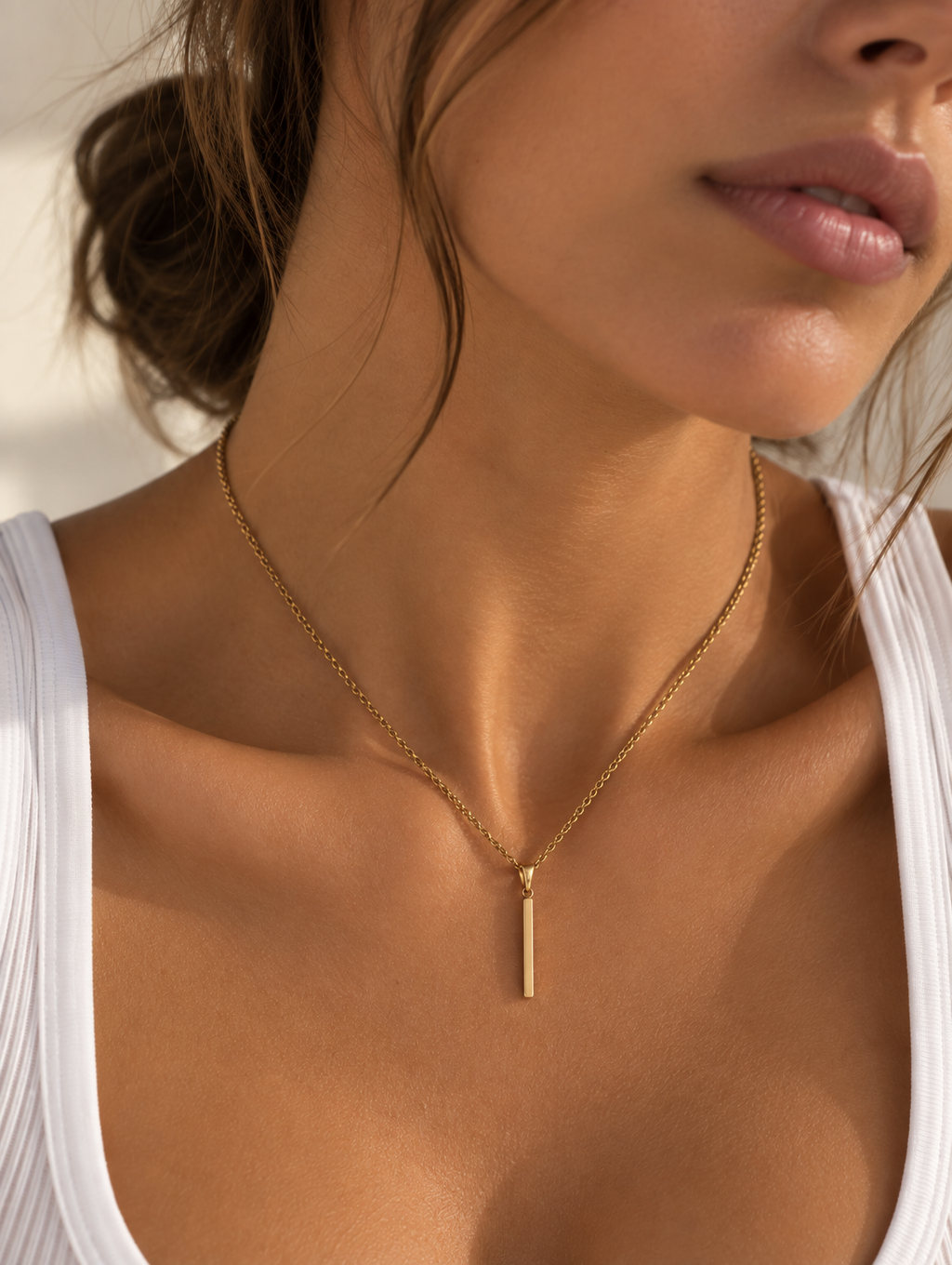 A close-up of a woman wearing a delicate gold chain necklace with a slim vertical bar pendant, styled with a white ribbed tank top.