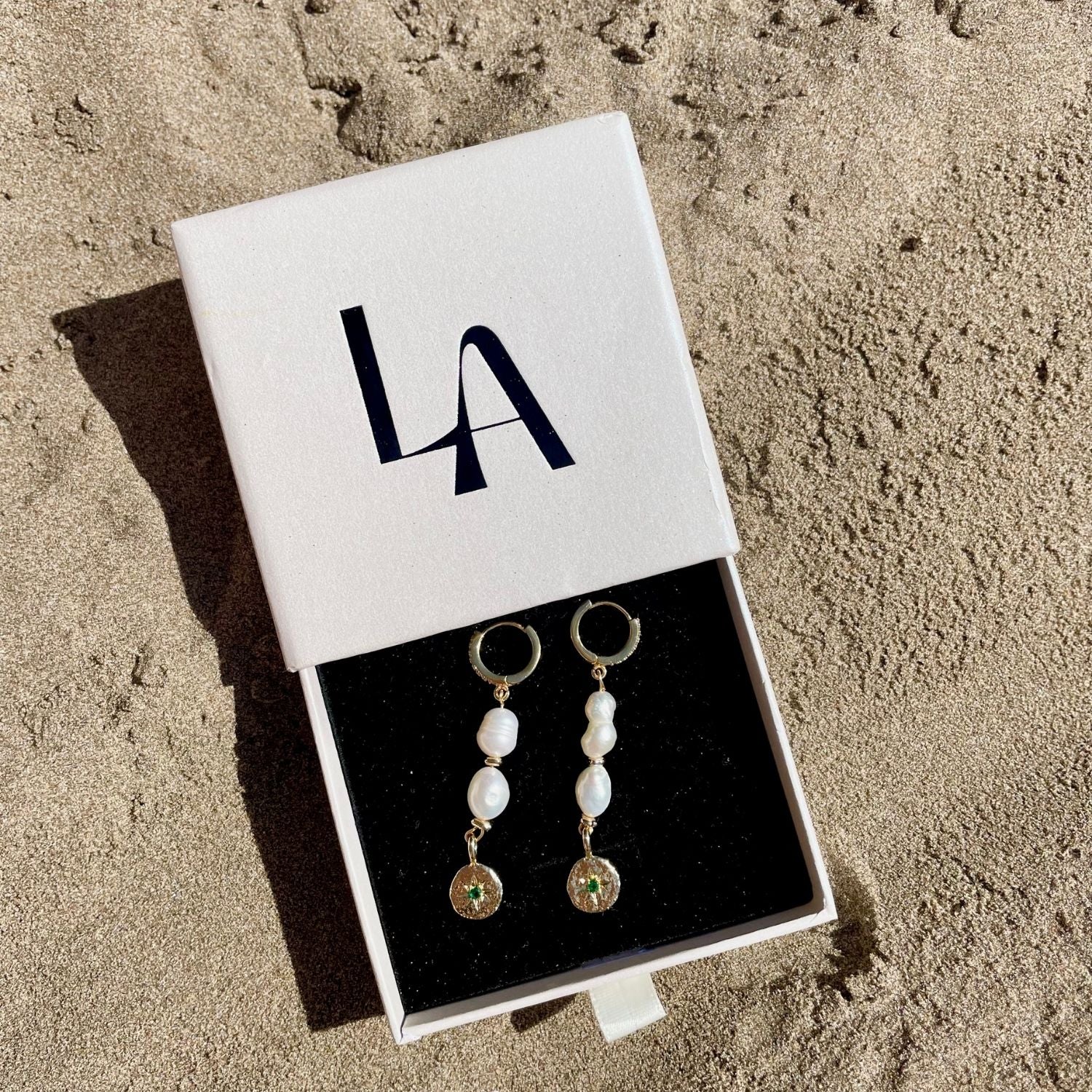 Pair of pearl earrings in a box with 'LA' branding on a sandy background