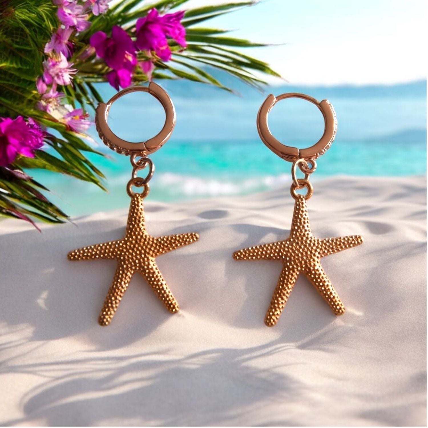 Gold starfish earrings on a sandy beach with pink flowers and blue sky in the background