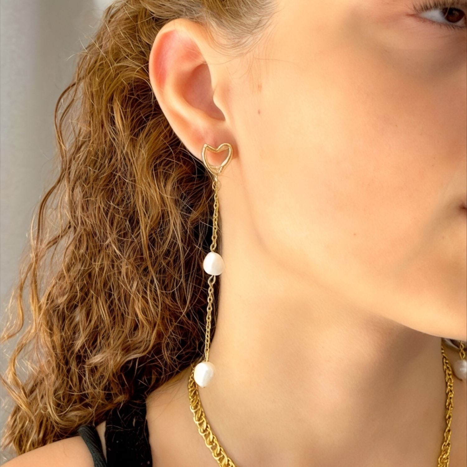 Close-up of a person wearing a gold chain earring with heart and pearl details.