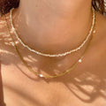 Two necklaces, one gold and one with white beads, on a person's neck against a neutral background.