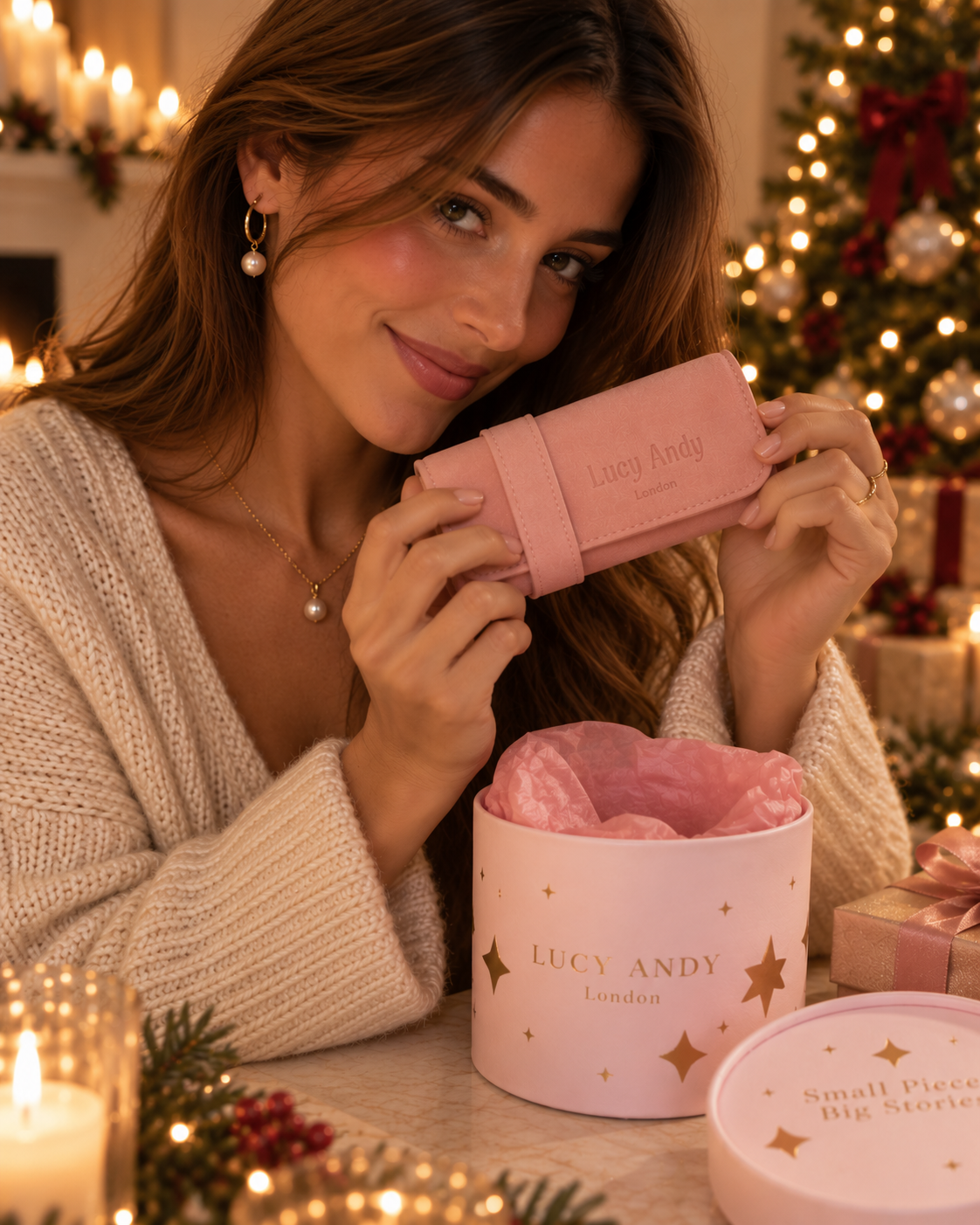 A woman in a cozy knit sweater holds a light pink rolled travel bag with 'Lucy Andy london' embossed on it, positioned above an open pink gift box with gold stars and the brand name 'LUCY ANDY London' printed on it. The background is warmly lit with festive Christmas lights.