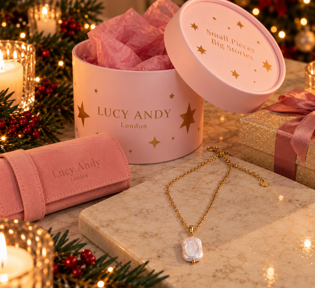 A pink round gift box with the lid open, filled with pink tissue paper, alongside matching pink jewelry packaging and gold necklace with white stone pendants on a marble surface. The box is branded LUCY ANDY London with gold star accents, and the scene is styled with candles, pine branches, and warm holiday lights.