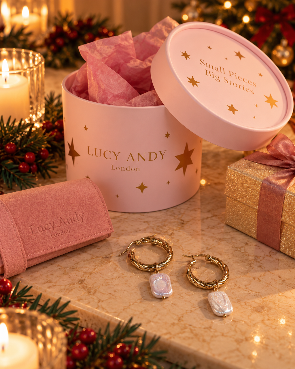 A pink round gift box with the lid open, filled with pink tissue paper, alongside matching pink jewelry packaging and gold hoop earrings with white stone pendants on a marble surface. The box is branded LUCY ANDY London with gold star accents, and the scene is styled with candles, pine branches, and warm holiday lights.