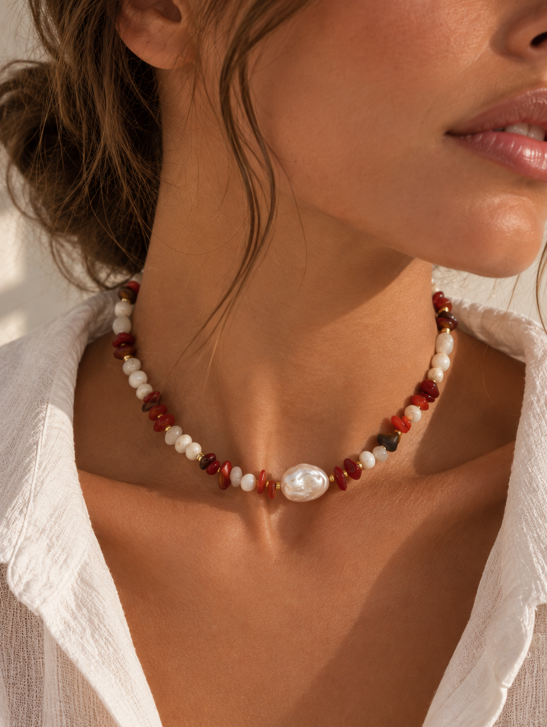 A woman wearing a bohemian-style beaded necklace with white pearls, red agate beads, and a central white pearl, styled with a white linen shirt.