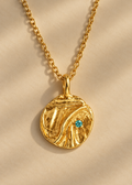 A textured gold pendant necklace with a small turquoise gemstone, displayed on a soft beige fabric background with elegant shadows.